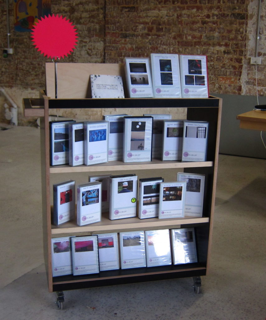 A picture of a library trolley filled with DVD and VHS covers, sitting in the middle of an industrial looking space, with tables and chairs on either side.