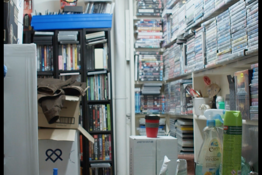 A behind the scenes view of a video shop store room, with shelving almost to the ceiling full of DVDs and assorted storage boxes, cleaning products and coffee mugs amongst them.